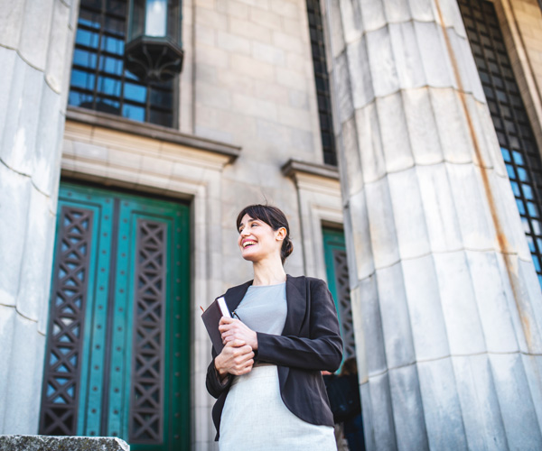 Low angle view of Hispanic female student in mid 30s looking away from camera while standing at entrance to Neo-Classical university building with book.